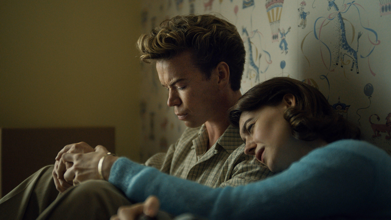 A man and woman dressed in clothing from the 1950s sitting down against a wallpapered wall and the woman has her head on the man's left shoulder