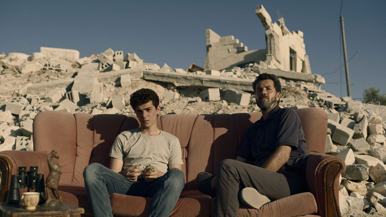 A younger man and an older man sitting on a brown couch amidst rubble