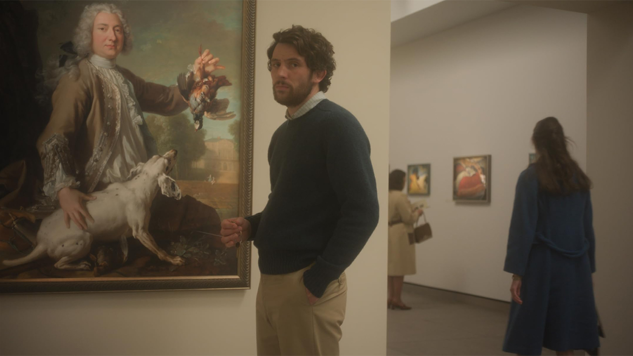 Young man with curly brown hair in museum with portrait behind him